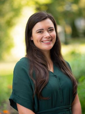 Victoria smiles, wearing a dark green dress shirt. She stands in front of a sunny green outdoor scene